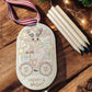 Wooden laser engraved ornament with outline of reindeer bicycling. Colored pencils stacked in the background.